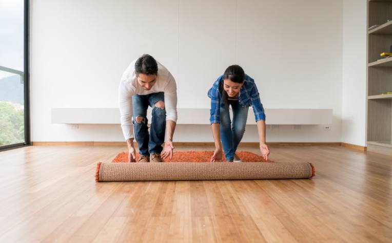 two people unrolling orange carpet in room with wood looking flooring and large windows.